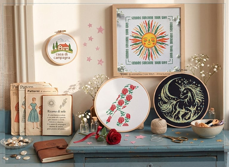 Bordados de punto de cruz Artmishka sobre una mesa vintage: sol sonriente, inicial floral y unicornio, entre patrones y flores silvestres.